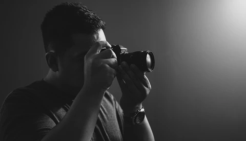 Photographer wearing glasses holding a DSLR camera up to his face in profile against a dark background with dramatic lighting.