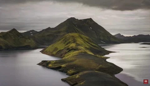 Moss-covered volcanic peaks rising from still water under overcast sky in Iceland.