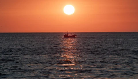 Silhouetted fishing vessel on calm water beneath a large sun during golden hour sunset.