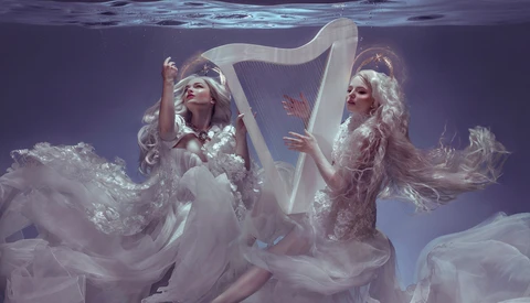 Two figures in flowing white gowns underwater with a harp, shot against purple-toned lighting.