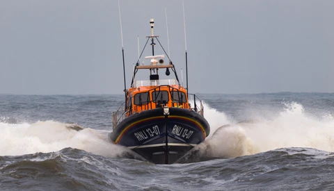 Orange and blue lifeboat navigating through large white-capped waves in rough ocean conditions.