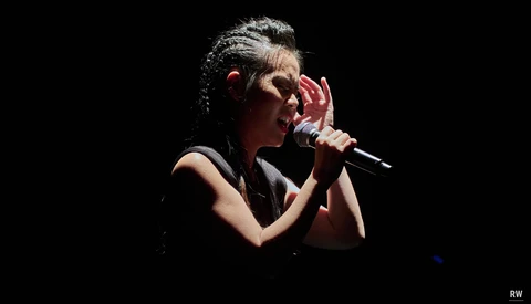 Female vocalist performing with microphone against dark background, wearing black sleeveless top.