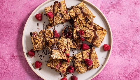 Golden granola bars with chocolate drizzle and fresh raspberries on a white plate against a pink textured background.