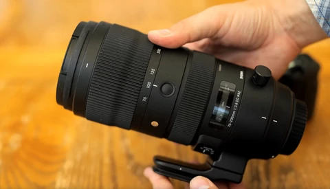Hands holding a black telephoto lens with visible focal length markings and focus ring against a warm wooden background.