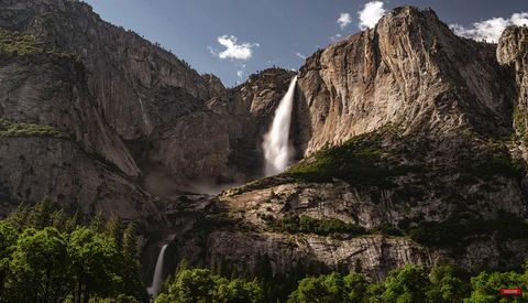 Tall waterfall cascading down sheer granite cliffs surrounded by dense green vegetation and rocky terrain.