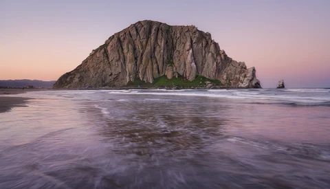 Massive rocky island with columnar basalt formations rising from shallow ocean waters at twilight.