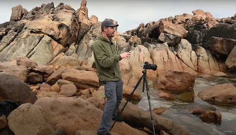 Photographer with camera on tripod standing among large granite boulders in rocky coastal landscape.