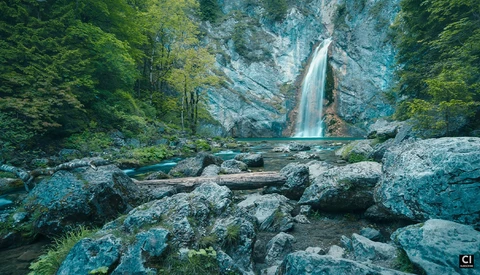 Waterfall cascading down a rocky cliff face surrounded by dense green forest and large boulders in foreground.