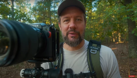 Videographer holding cinema camera with telephoto lens in forest setting during golden hour.