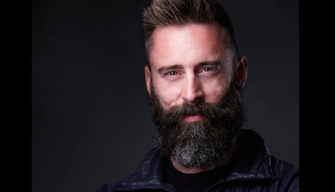 Man with full dark beard and styled dark hair wearing black jacket against dark background.