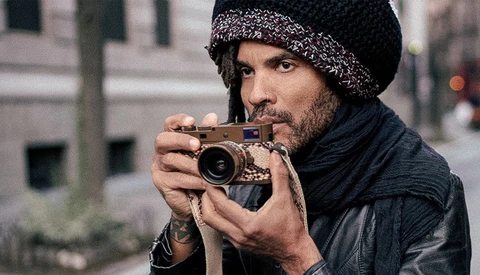 Bearded man in black beanie and leather jacket holding vintage rangefinder camera to his face.