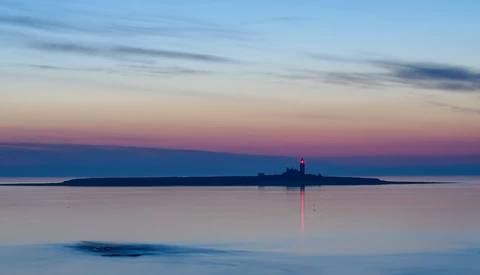 Lighthouse beam reflects across still water at twilight, with distant forested shoreline silhouetted against purple and blue sky.