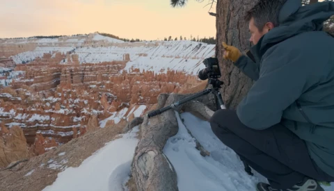 Photographer using a telephoto lens on a tripod at the edge of a snowy cliff overlooking a vast canyon landscape.
