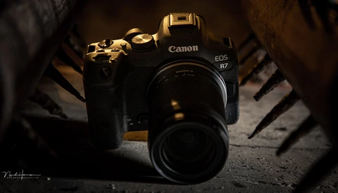 Canon EOS R7 mirrorless camera with attached lens on textured surface with dramatic side lighting.