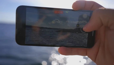 Hand holding smartphone recording video of ocean and sky at sunset or sunrise.