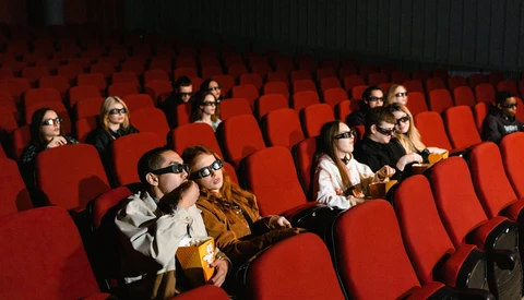 Audience members seated in a movie theater with red seats, some wearing glasses and holding popcorn.