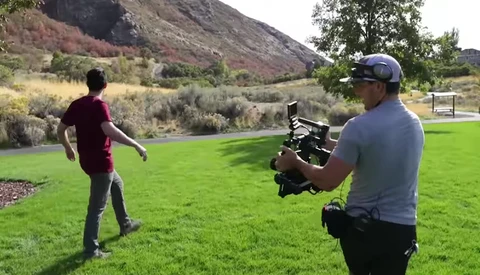 Cameraman operating a handheld cinema camera while filming a person in an open field with mountains and trees in the background.