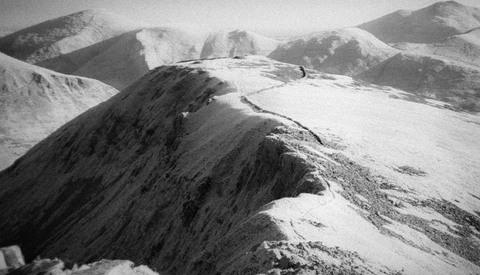 Snow-covered mountain ridge with exposed dark rock face under overcast alpine sky.