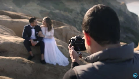 Wedding photographer shooting bride and groom posing on rocky terrain during golden hour.