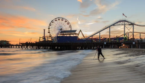 Surfer walking into the ocean at sunset with an amusement park pier and roller coaster in the background.