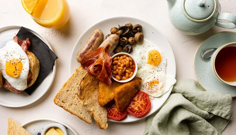 Overhead flat lay of a full breakfast spread with eggs, bacon, toast, and fresh fruit on white plates.