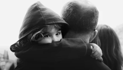 Child wearing a dark hoodie hugging an adult from behind in black and white.