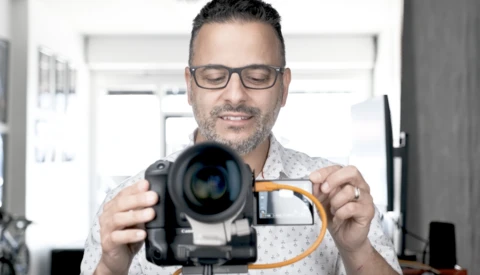 Photographer wearing glasses holding a video camera with an orange lens adapter attached.