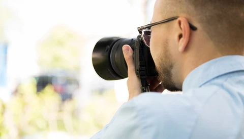 Photographer wearing glasses holding a camera up to his face while shooting outdoors.