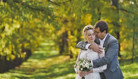 Groom embracing bride from behind in a tree-lined park setting, bride holding white flower bouquet.