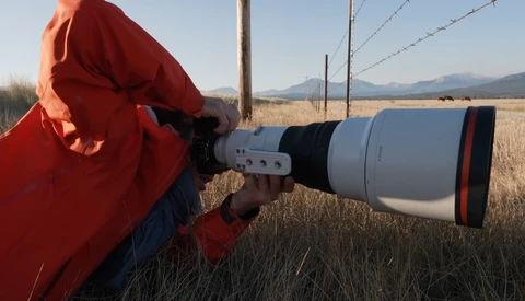 Photographer in red jacket using telephoto lens in open field with mountains in background.