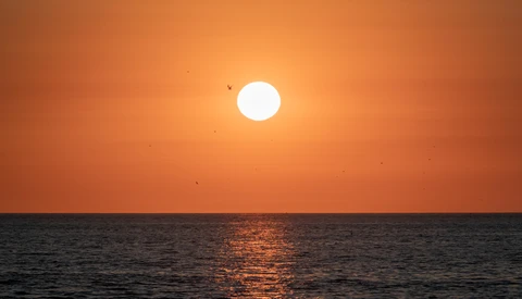 Bright sun positioned above a calm ocean horizon at sunset, casting a golden reflection across the water.