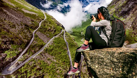 Photographer on rocky outcrop capturing winding mountain road through valley below with camera.