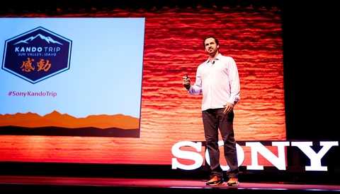 Speaker presenting on stage at a Sony event with red brick backdrop and illuminated signage.