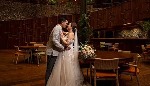 Bride and groom kiss in a modern wooden-walled venue with ambient lighting and floral arrangements.