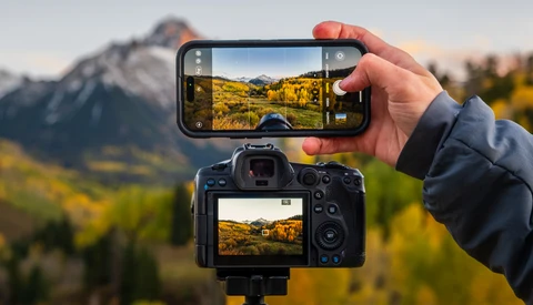 Smartphone and digital camera displaying identical mountain landscape scenes with golden vegetation and snow-capped peaks.