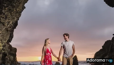 Couple holding hands on a beach at sunset, framed by large rock formations on either side.