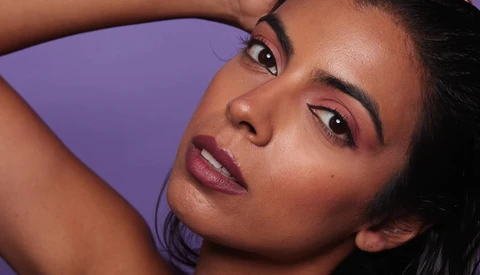 Woman with hand raised to head against purple background, wearing pink-toned makeup.