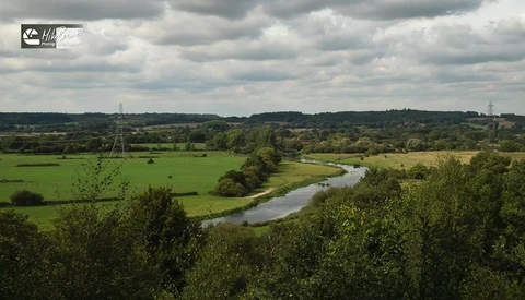 Meandering river cutting through pastoral English countryside with rolling hills, green fields, and distant woodlands under partly cloudy sky.