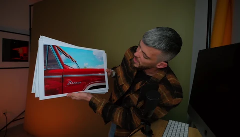 Photographer holding printed photograph of a red vintage car against a green wall in an office setting.