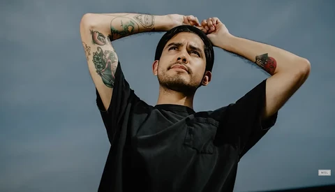 Tattooed man in black shirt with hands behind head against blue-gray sky background.