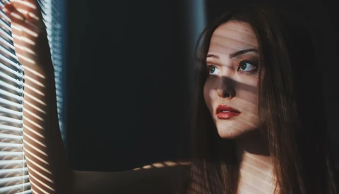 Woman with long dark hair looking toward a window with venetian blinds casting striped shadows across her face.