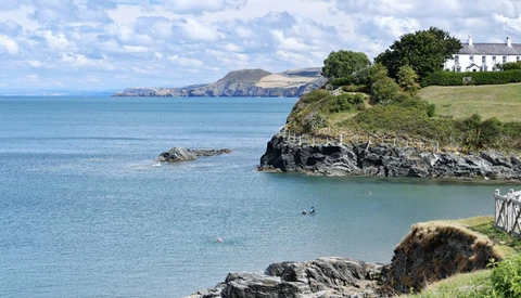 Turquoise coastal cove with rocky outcrops, white house on green headland, and distant cliffs across blue water.