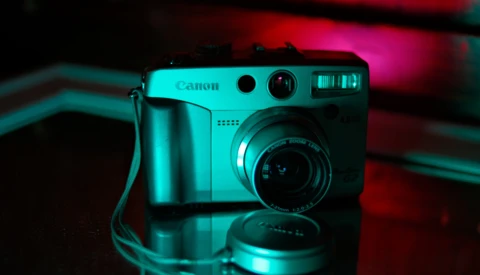 Canon compact camera with lens cap nearby, lit with cyan and magenta neon lighting against a dark background.