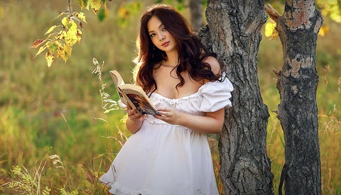 Woman in white off-shoulder dress holding dried corn stalks beside a weathered tree trunk.
