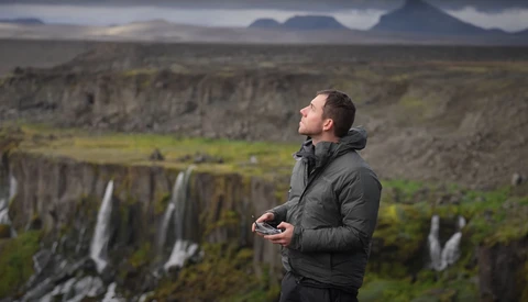 Man holding a drone controller overlooking a vast landscape with waterfalls and basalt cliffs.