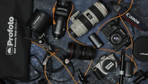 Professional photography gear laid flat on a textured dark surface, including camera bodies, lenses, lighting equipment, and cables with orange straps.