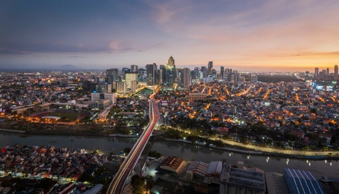 Cityscape at dusk with illuminated skyscrapers, a river, and light trails from traffic on an elevated highway.