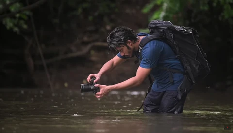 Photographer wading in shallow water while operating a camera, wearing a blue shirt and black backpack.