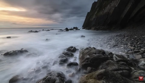 Rocky coastline at dusk with smooth water flowing around dark boulders beneath a cliff.