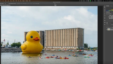 Giant yellow rubber duck floating on water with industrial grain elevator and kayakers in background.